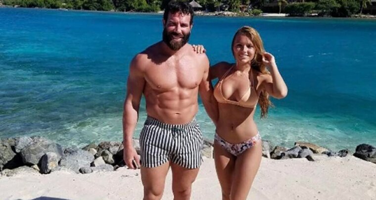Dan Bilzerian in striped black-and-white pinstripe swim trunks and a woman in a pink bikini standing on a beach in front of the ocean.